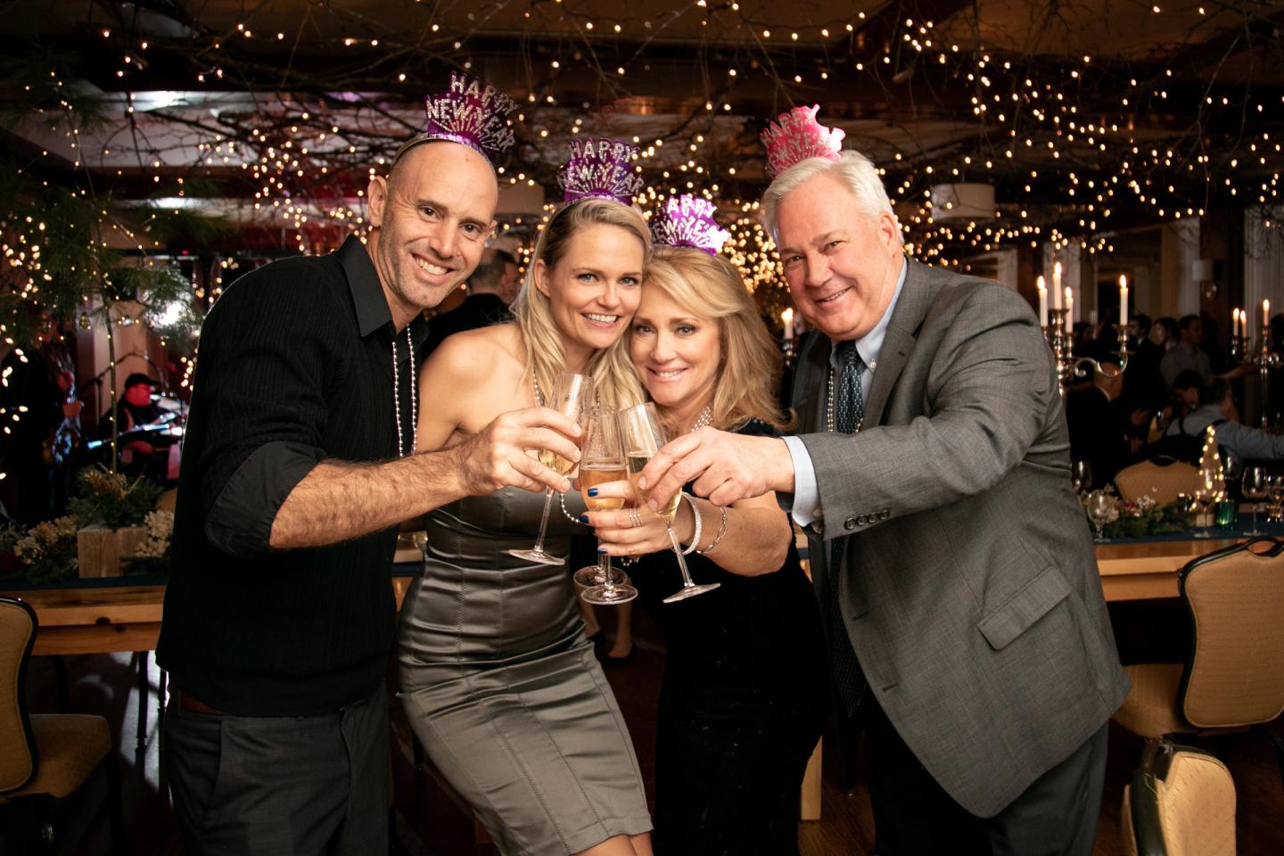 4 well dressed people toasting champagne glasses. twinkle lights and new year's eve party hats are in the background