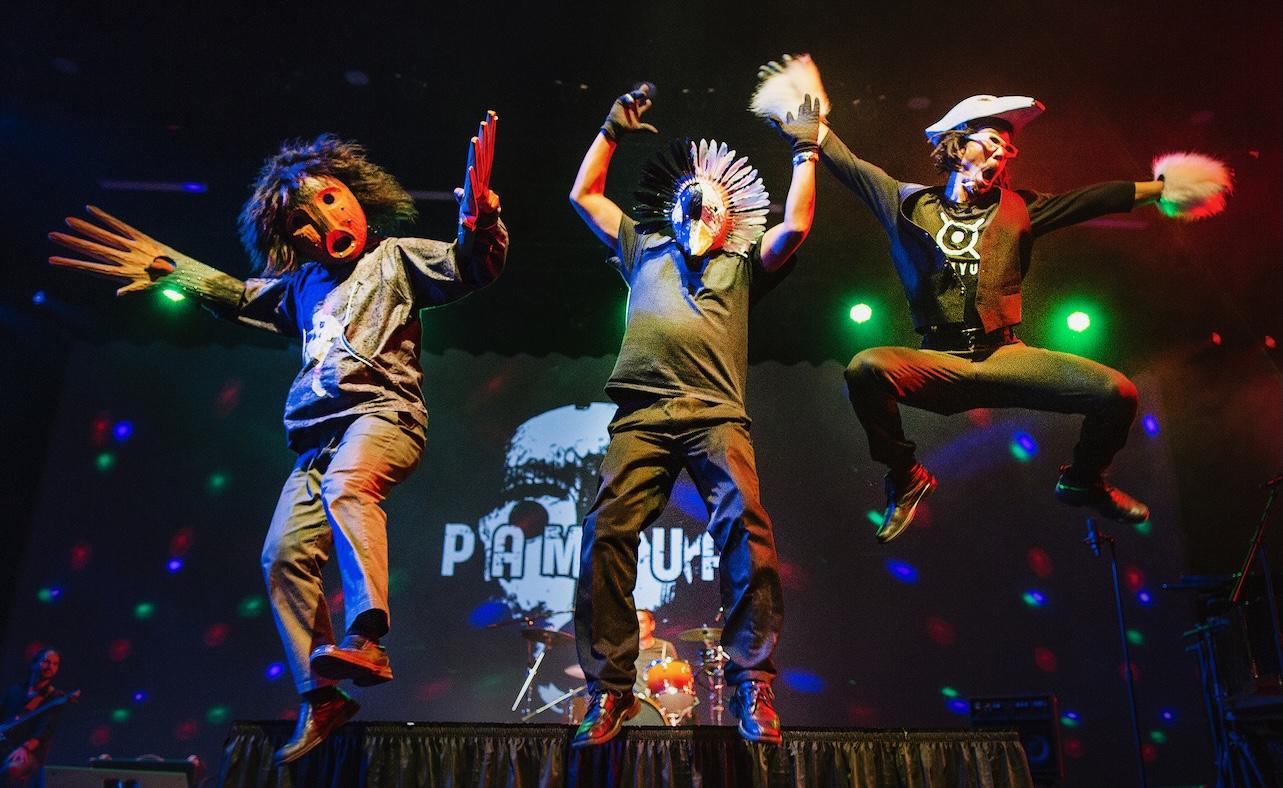 Three band members jump in the air wearing Indigenous Alaskan masks and gloves.