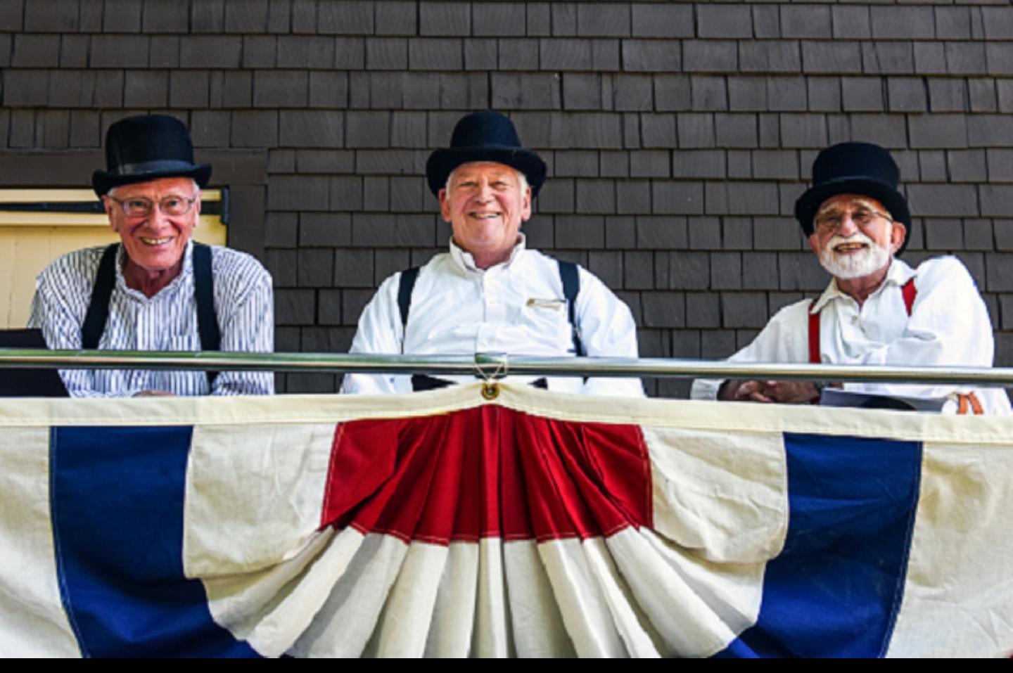 Three men dressed in old timey outfits sitting behind a red, white, and blue flag.