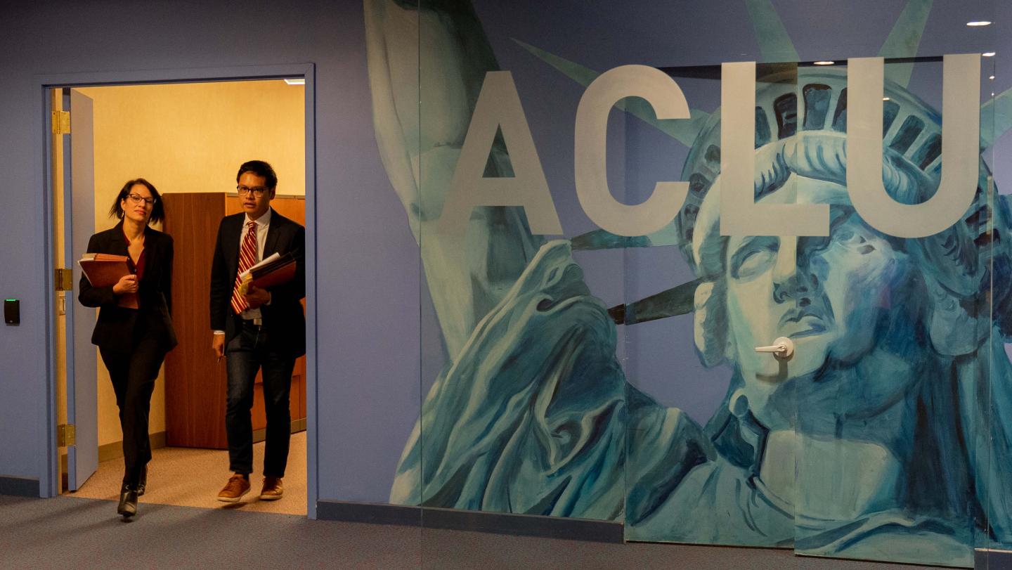 Members of the ACLU stride through doorway by wall, which reads "ACLU"