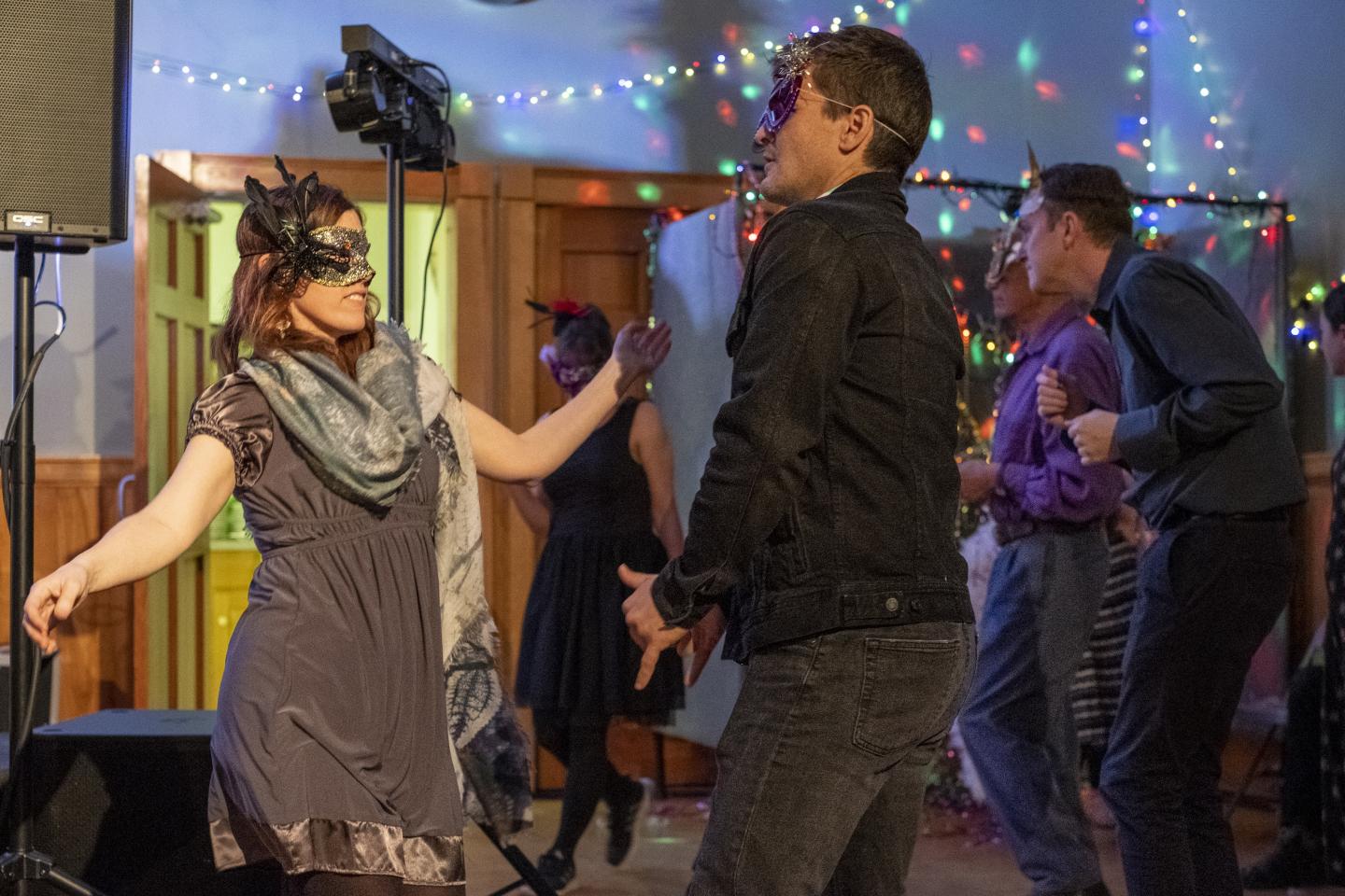 Two people dance while wearing masquerade masks. Others do the same in the background. There are string lights along the walls.