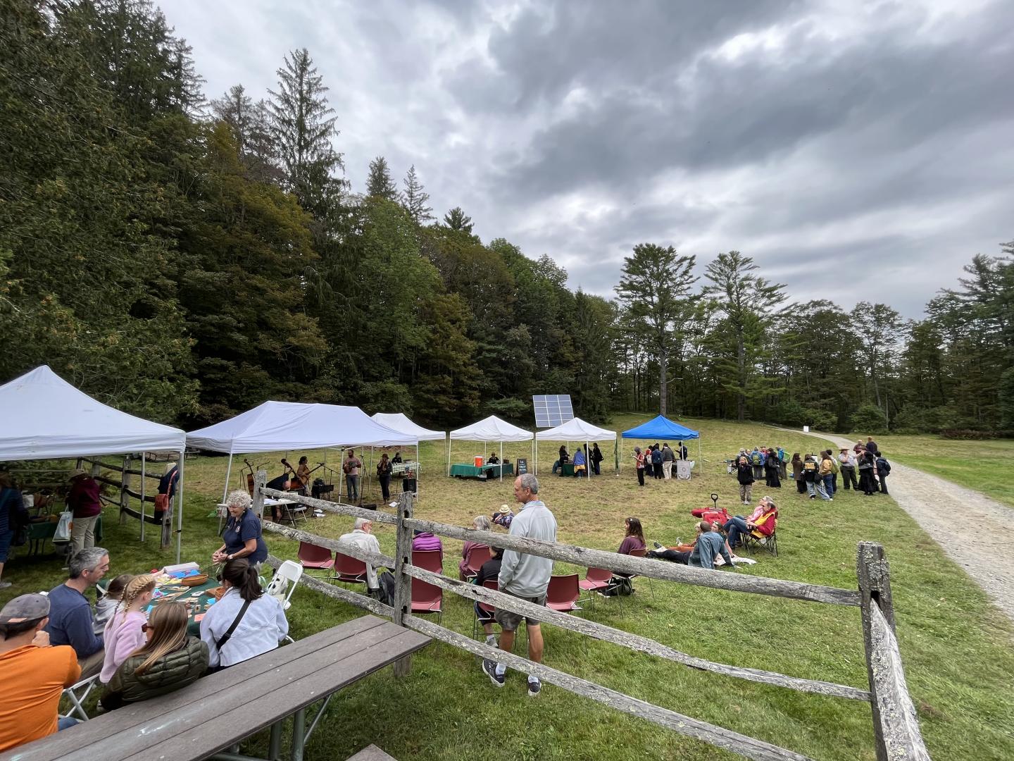 wide shot of field with picnic table and wood fence in foreground and 5 pop up tents in background, people watching live music sitting on chairs and blankets