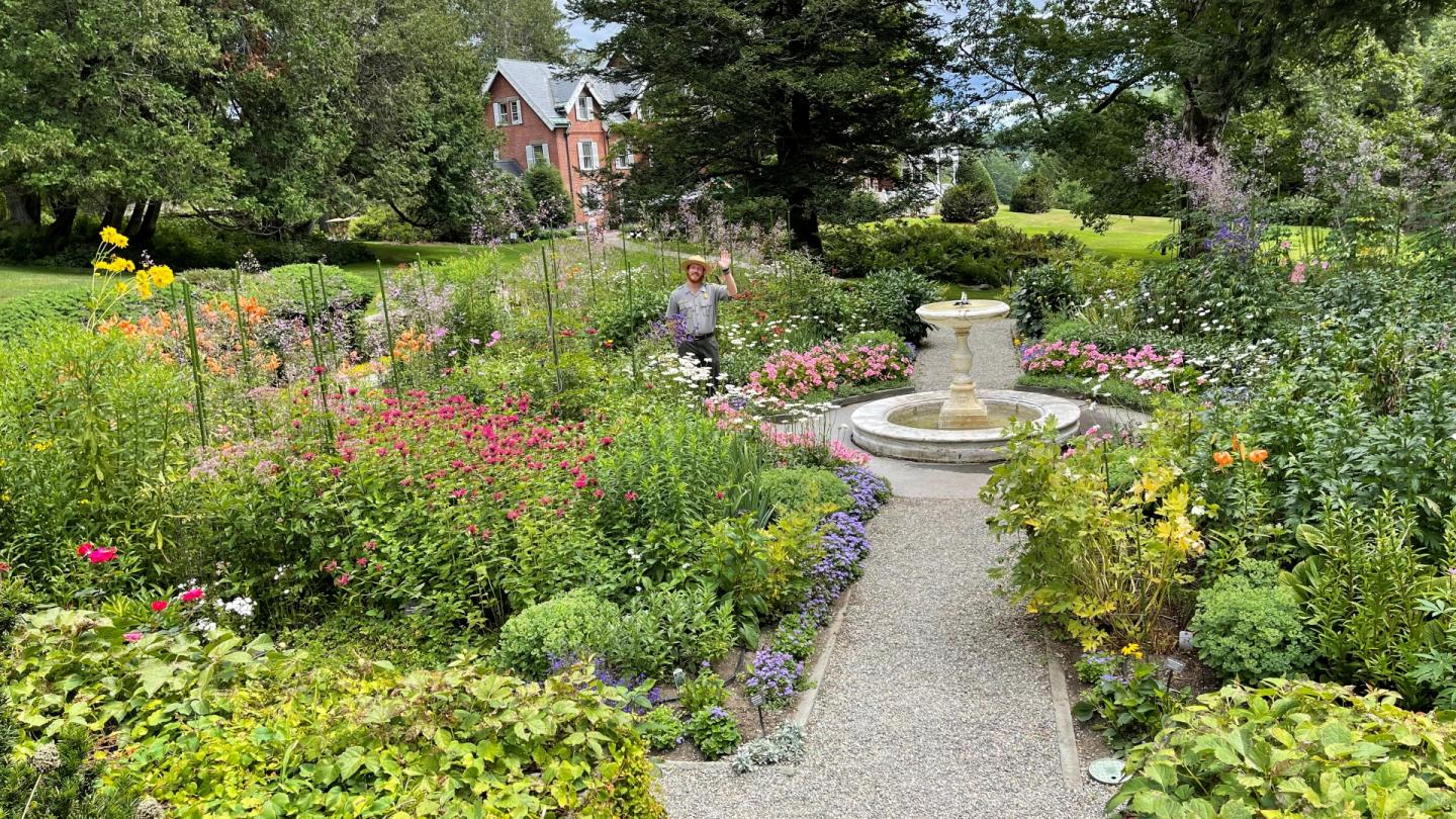 Park Ranger stands and waves at camera in formal four square garden with large historic home in background