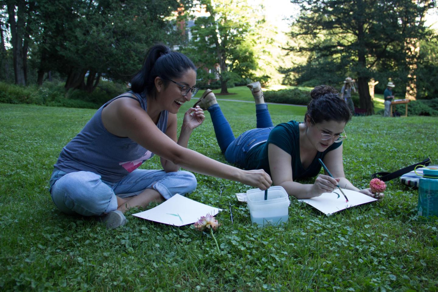 Two women laugh while sitting in the grass and painting