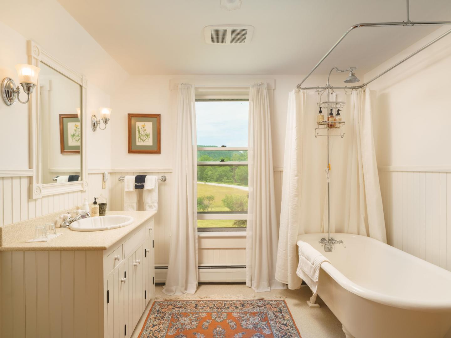 A vintage-style bathroom with a clawfoot tub and shower, cream vanity, large mirror, and a window overlooking the Vermont countryside at The Farmhouse Inn.