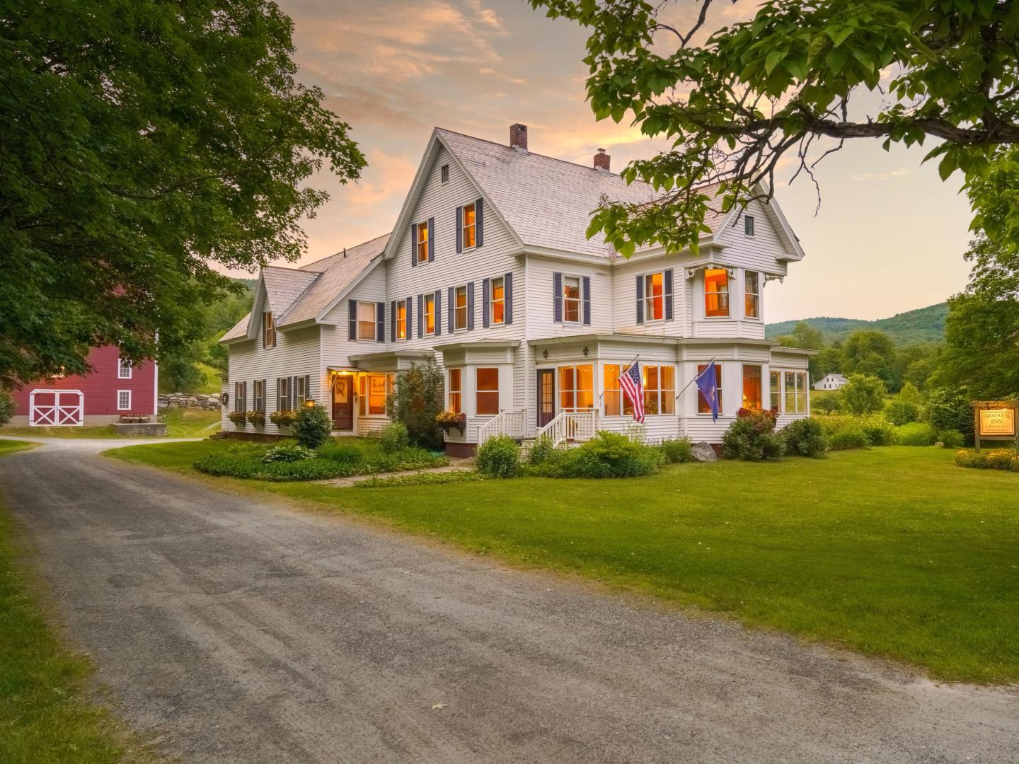 A large white farmhouse with glowing windows at sunset, surrounded by green lawns and trees, with a red barn in the background and American flags on the front porch.