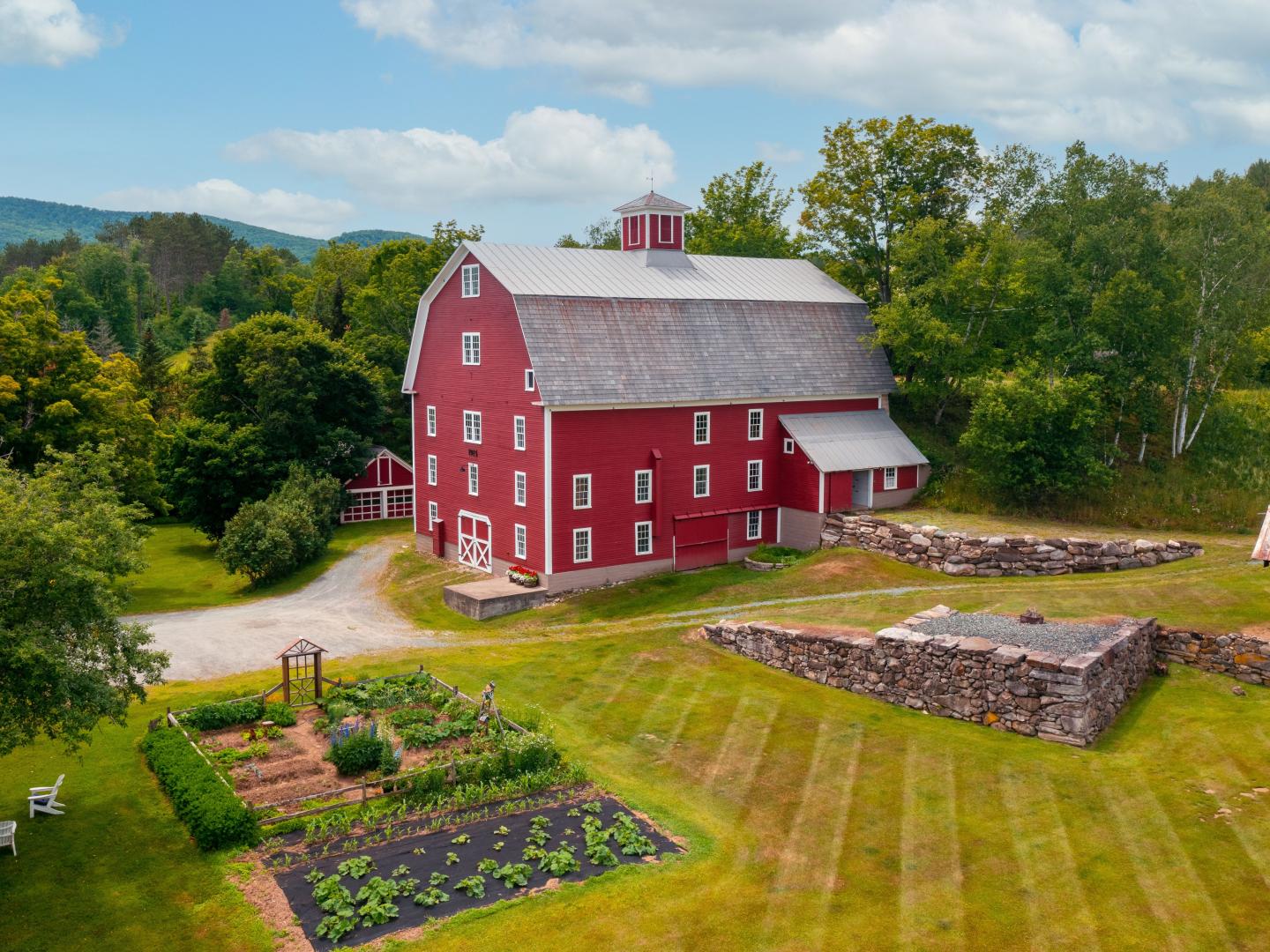 A bright red barn at The Farmhouse Inn, surrounded by green trees and stone walls, with a lush vegetable garden in the foreground and mountains in the distance.