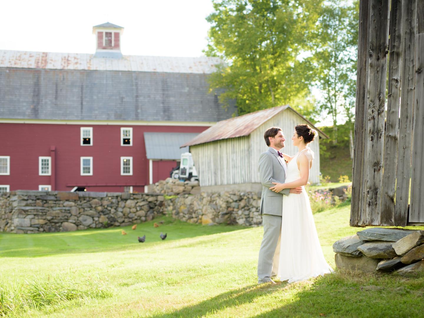 A bride and groom smiling at each other in front of a rustic barn at The Farmhouse Inn, with chickens roaming the grass and sunlight filtering through the trees.