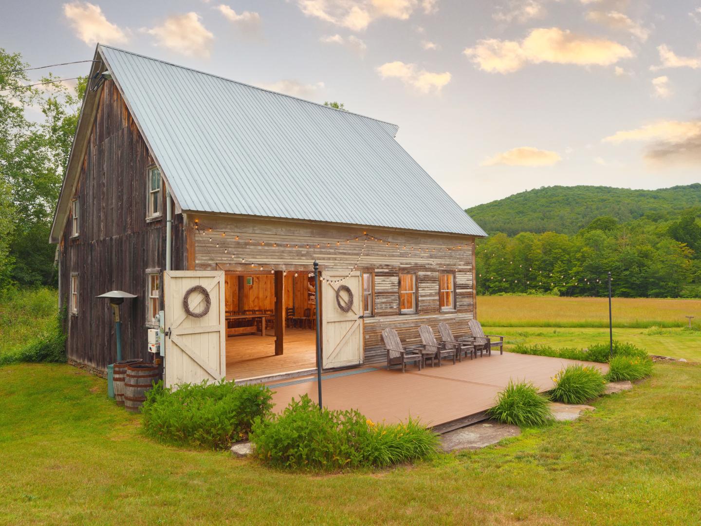A rustic wood barn with open doors, string lights, and Adirondack chairs on a deck, set against a backdrop of open fields and forested hills at The Farmhouse Inn.