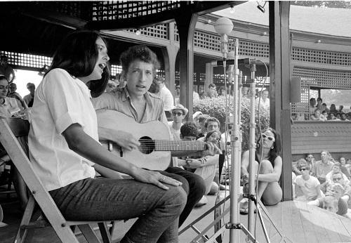 Black and white photo of a female and male singer on a stage with a guitar. Joan Baez and Bob Dylan.