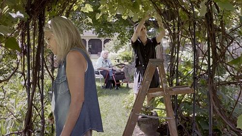 Female wearing a sleeveless blouse outdoors under an arbor with a man on a ladder and a person sitting in the background.