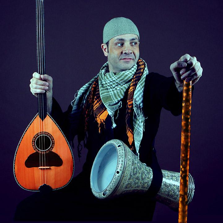 Photo of musician holding three instruments (something that looks like a guitar, a drum, and a hollow stick) and looking off to the left.