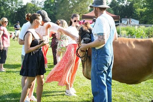Guests holding beverages whilst petting a cow with farmer in overalls.