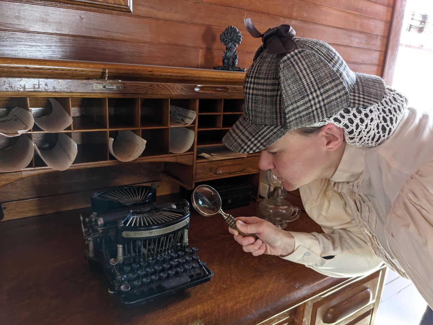A person in a Deerstalker hat examining a typewriter with a magnifying glass