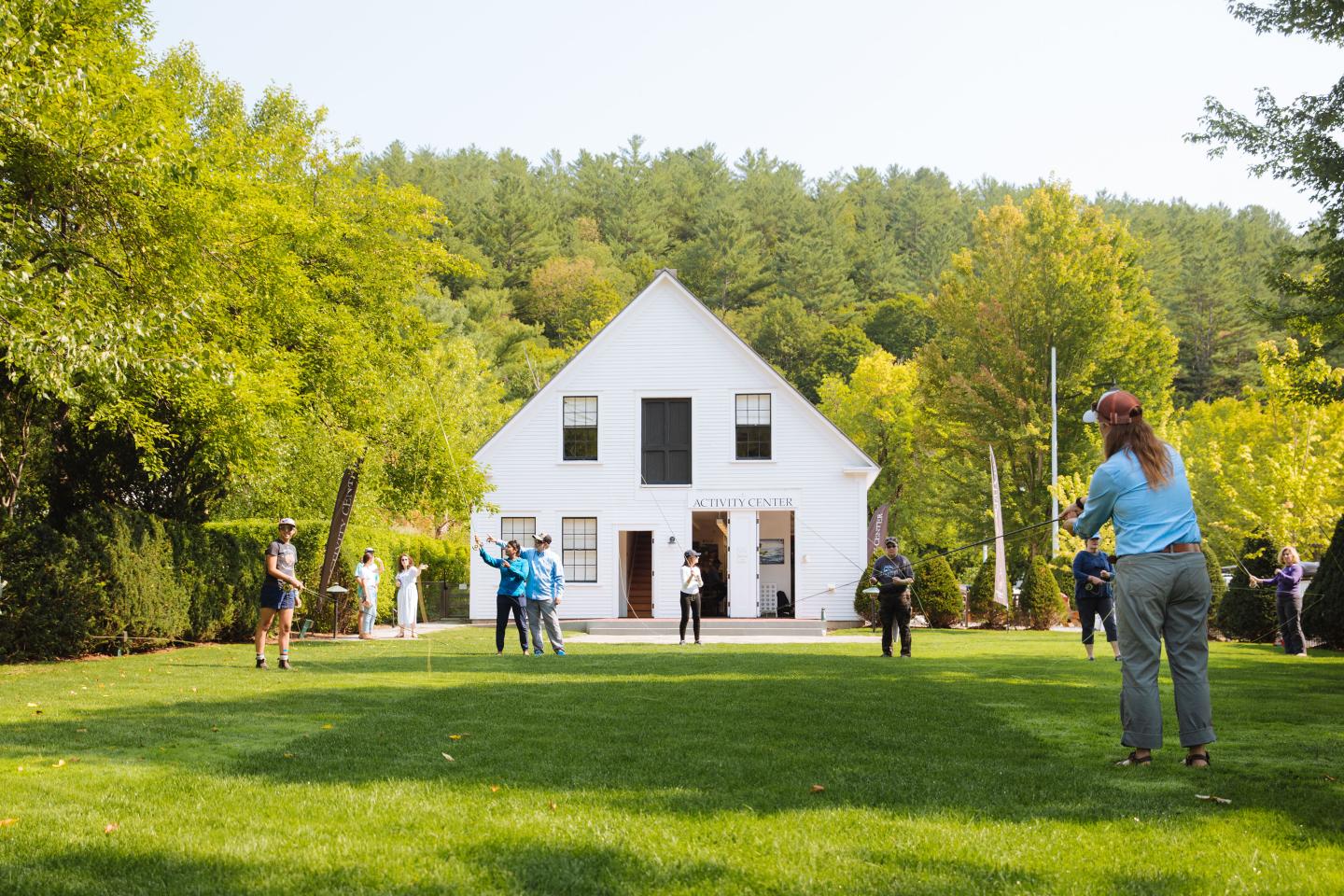 A group learning to fly fish on a green lawn with a white activity center barn in the background