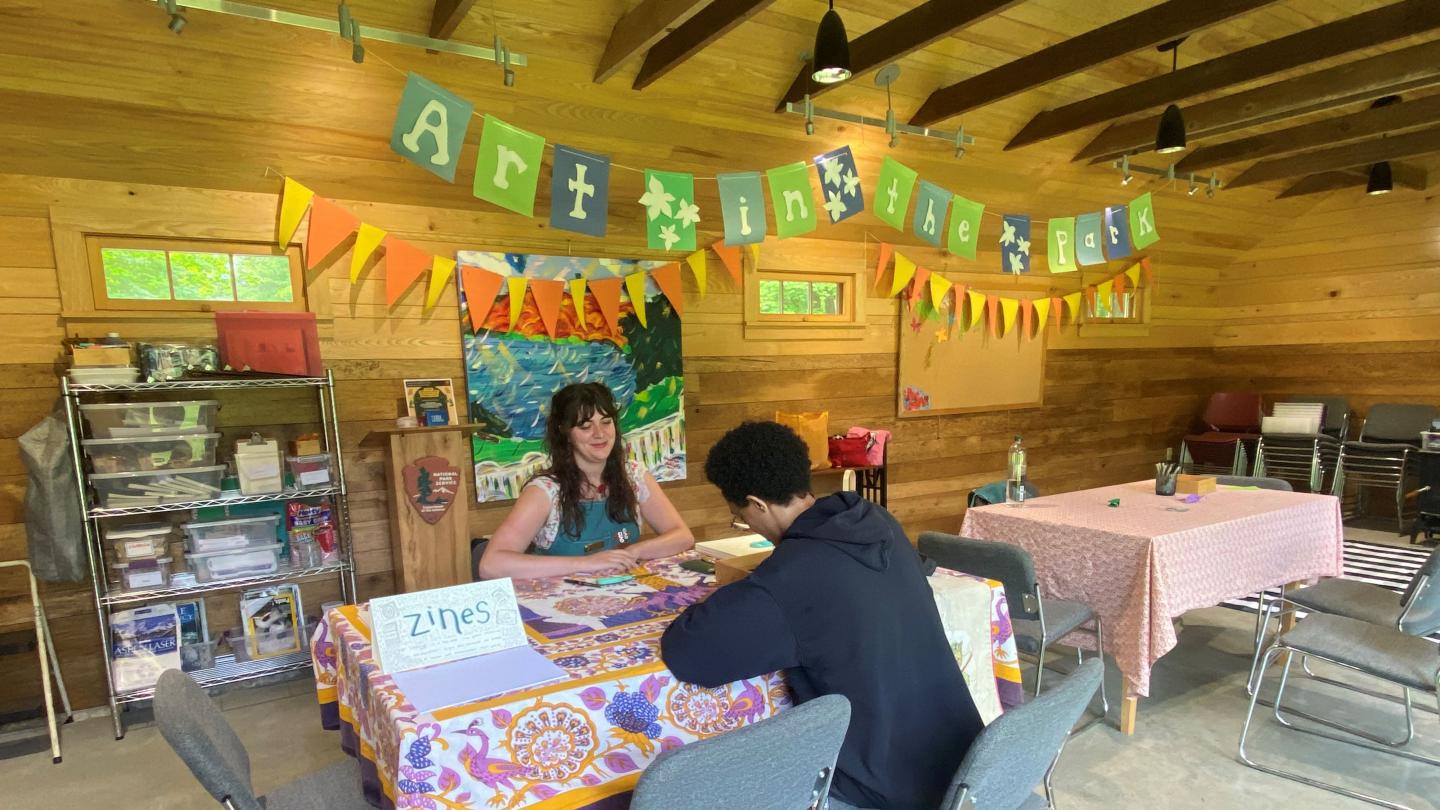 two people sit at a table that has a sign that says "zines" sign behind them says "art in the park"