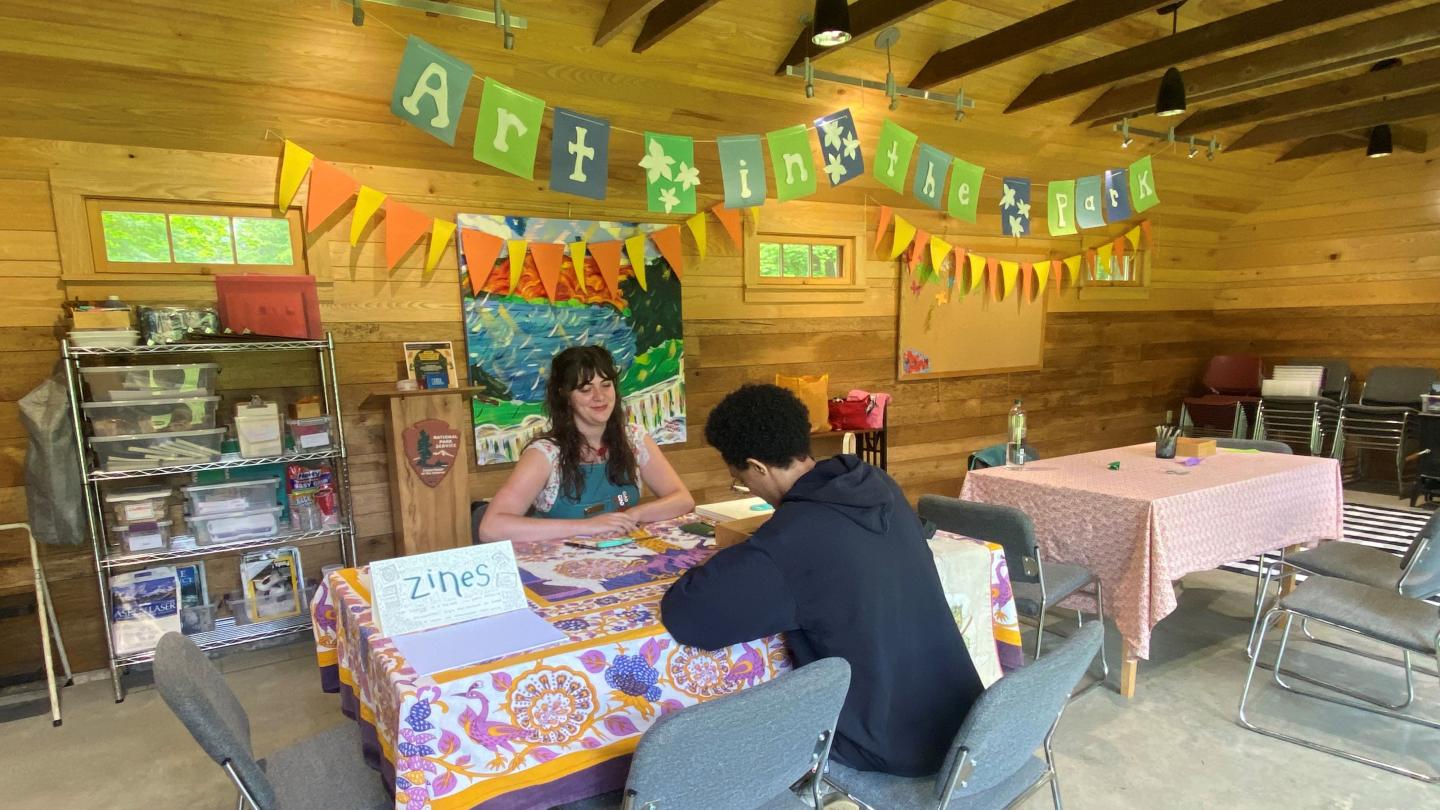 two people sit at table with sign that says art in the park in background