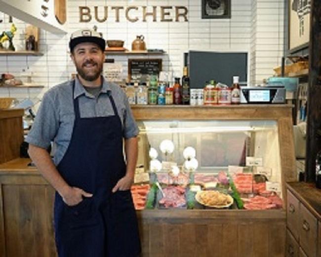 Various cuts of meat in a glass case with a man in an apron, ball cap, beard standing next to it.