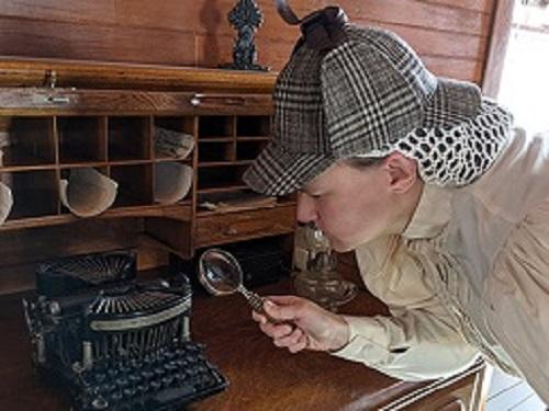 Person dressed in a Sherlock Holmes hat with magnifying glass looking at an old typewriter.