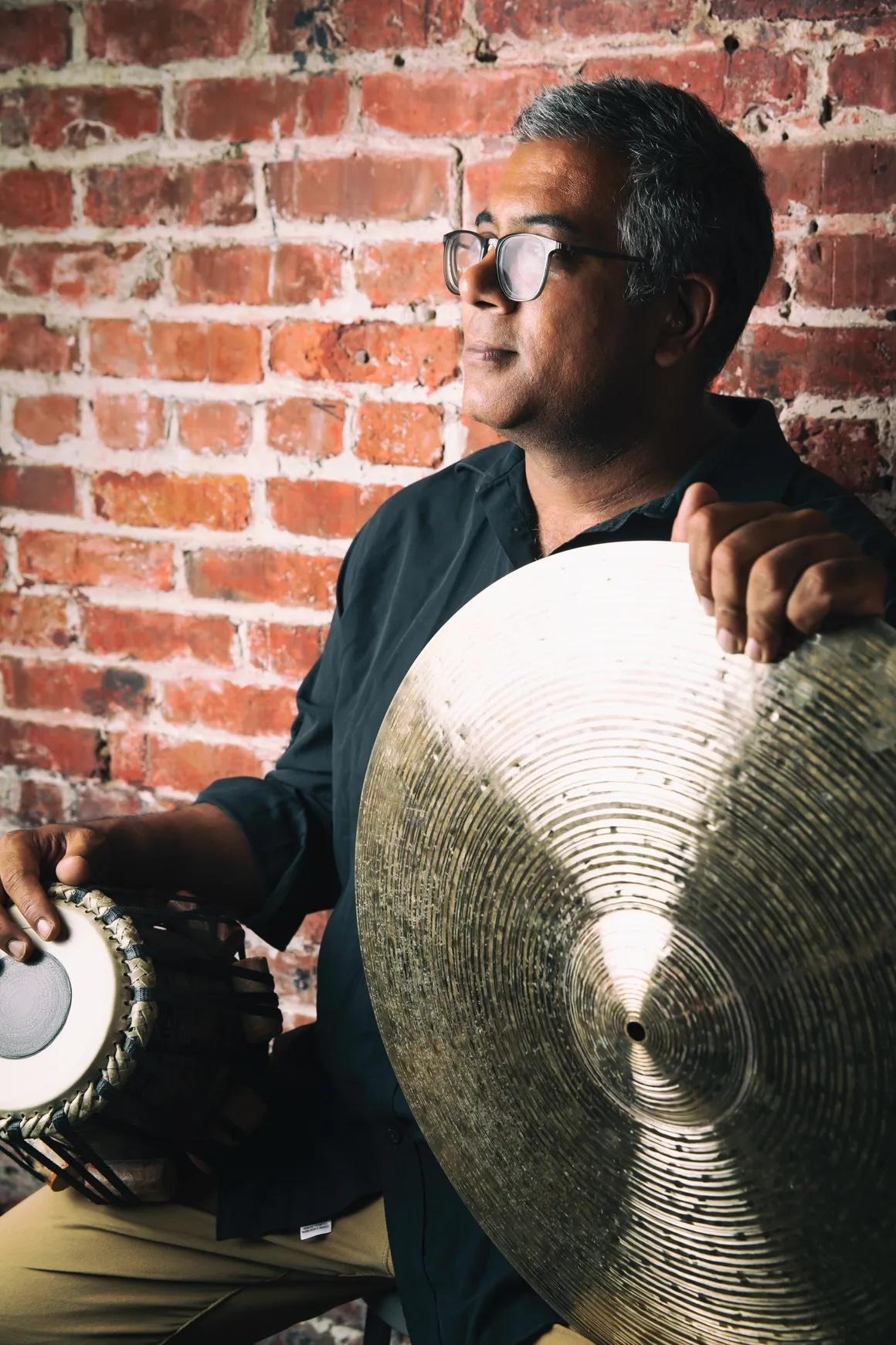 Sameer Gupta sits in front of a brick wall and looks off to the left. He holds two musical instruments, one in each hand. To the left is a tabla, a small Indian drum. To the right is a big metal circle, likely another percussion instrument.