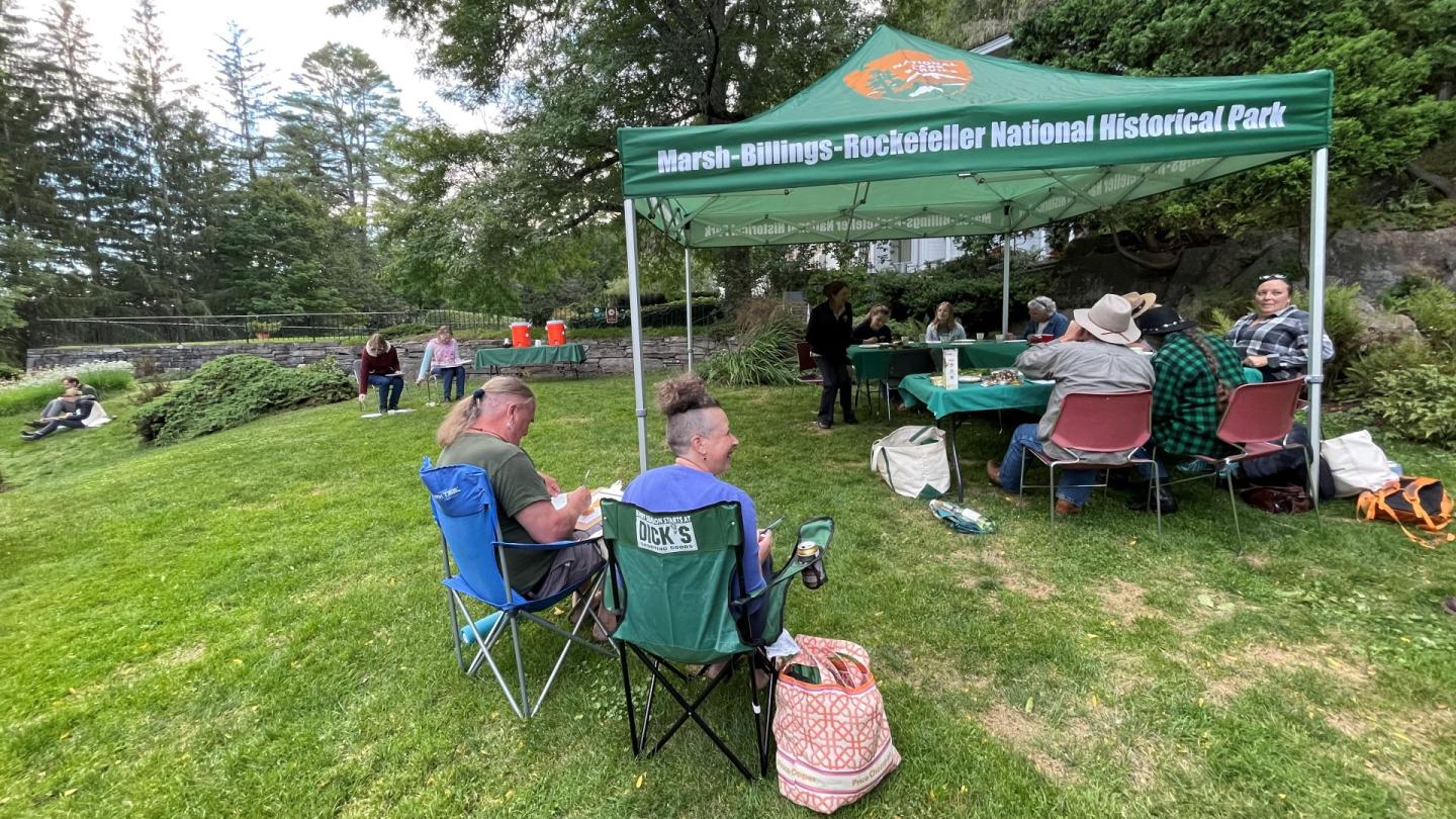 Group of people sit in chairs and paint on green lawn under tent that says "Marsh-Billings-Rockefeller National Historical Park)