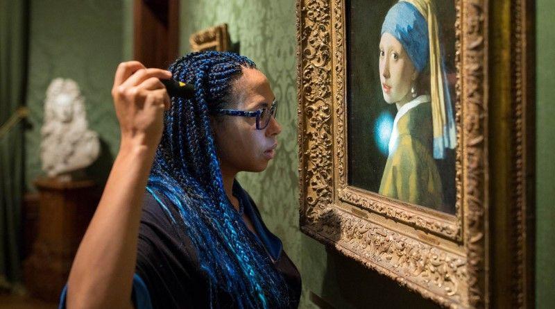 Woman with long hair dyed blue shining a small flashlight on a painting hanging on a wall.