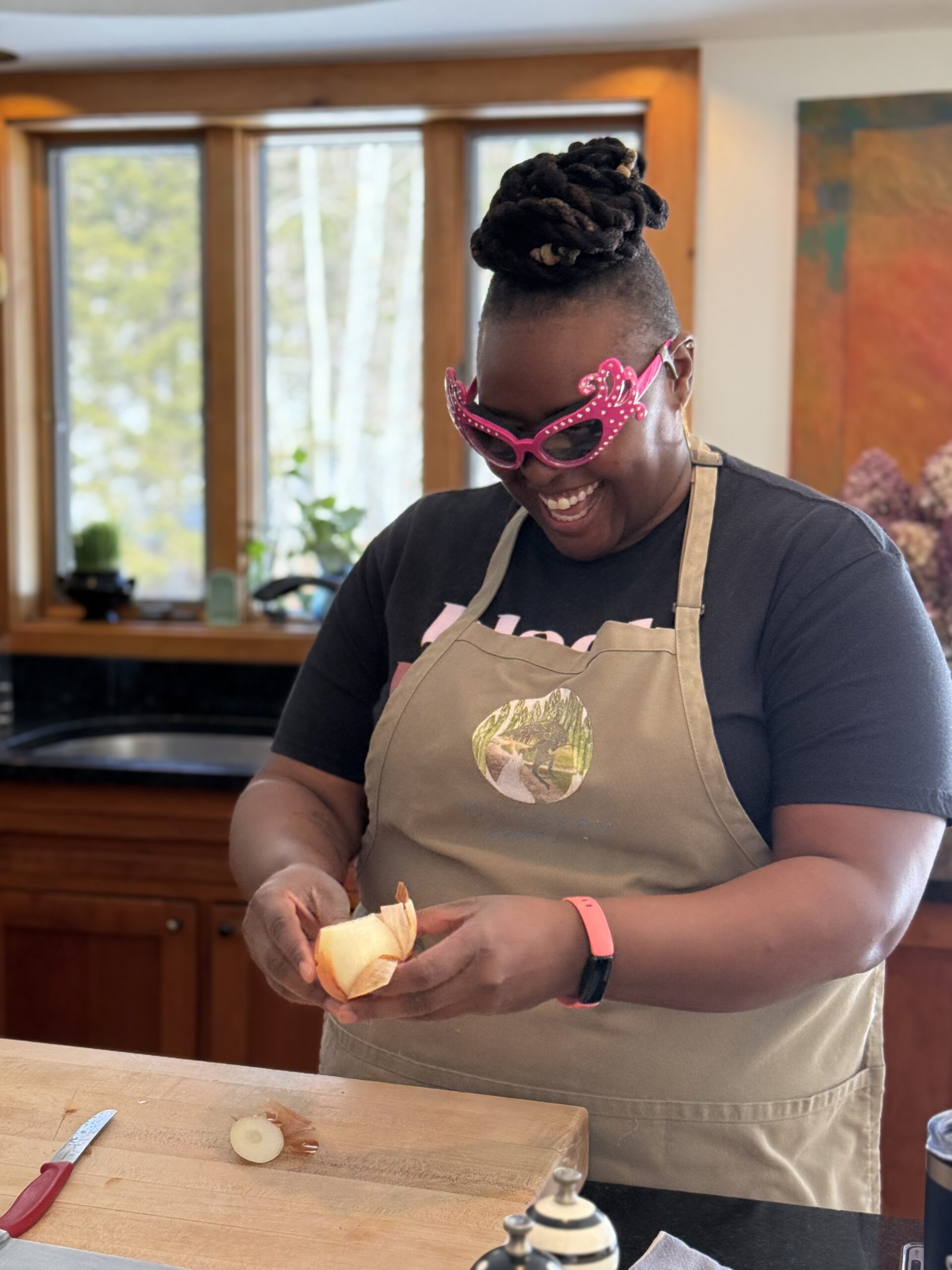 A woman wearing an apron and protective glasses smiles widely while slicing an onion