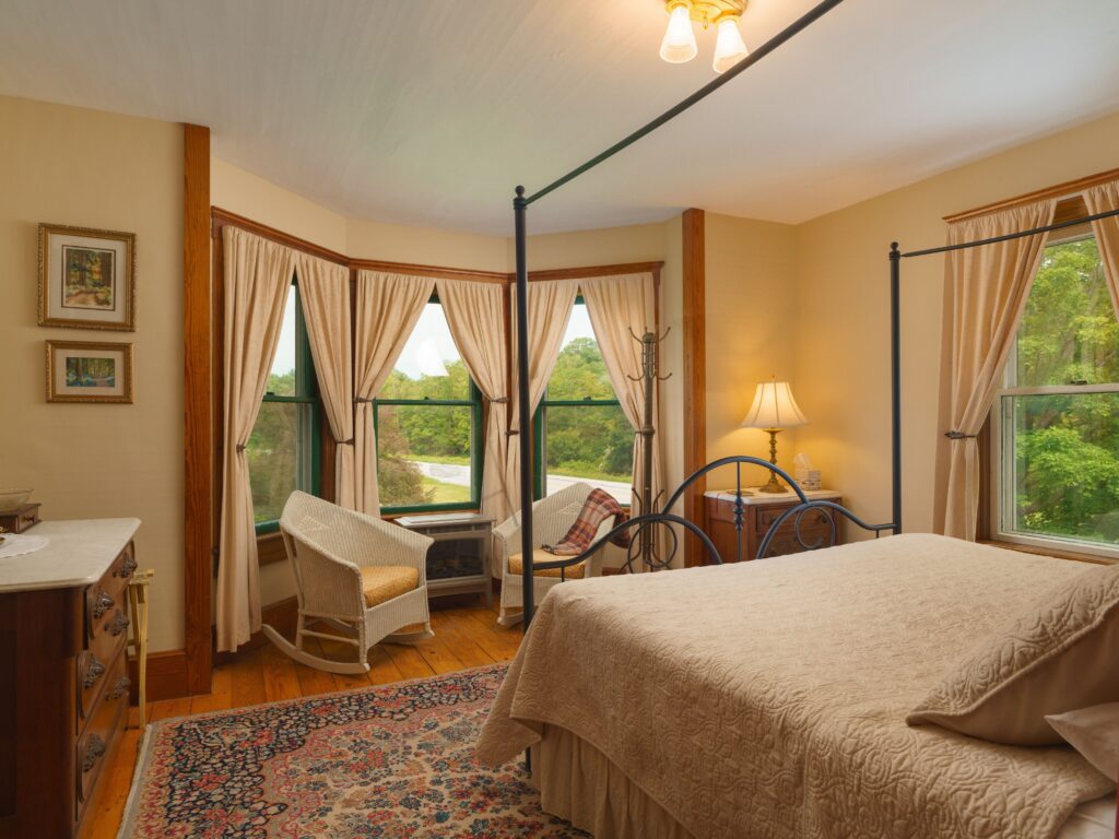A cozy guest room at The Farmhouse Inn featuring a four-poster bed, vintage-style furniture, wicker rocking chairs by large windows, and views of the surrounding greenery.