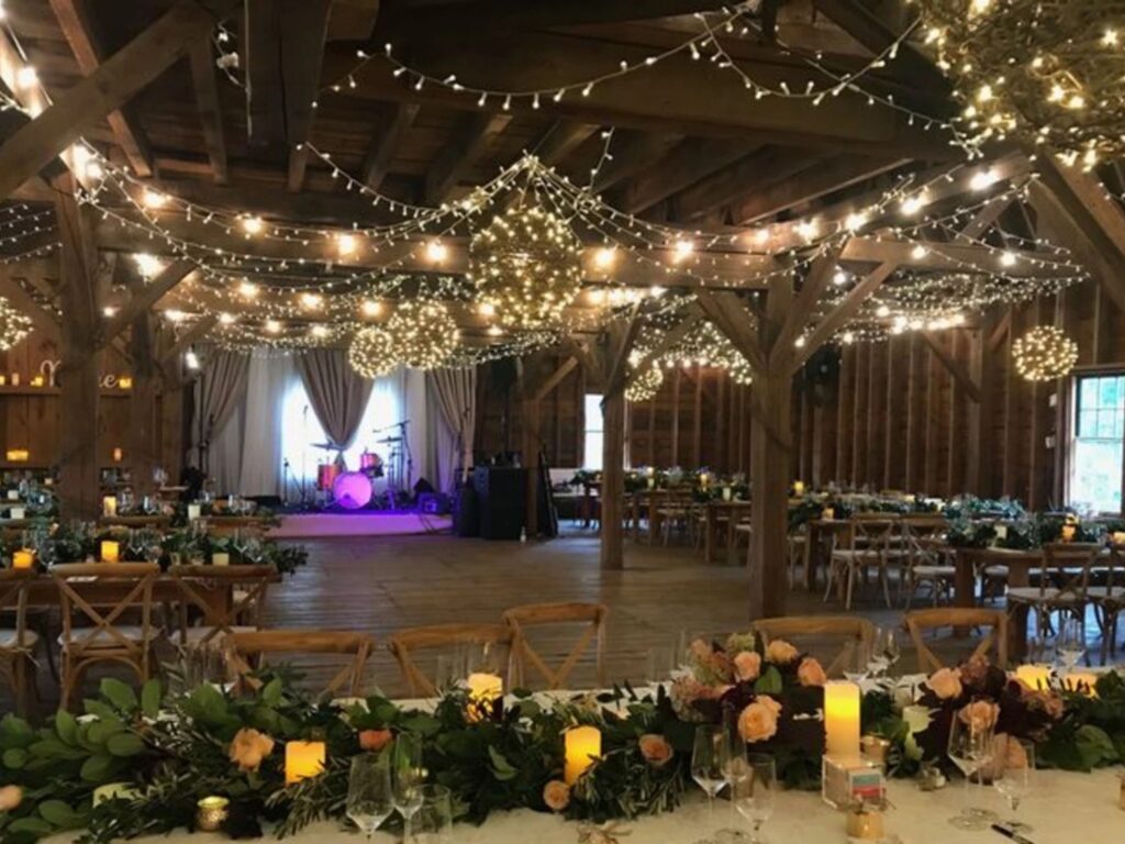 The interior of a rustic barn decorated for a wedding, with string lights overhead, floral centerpieces, candles, and a stage with musical instruments at The Farmhouse Inn.
