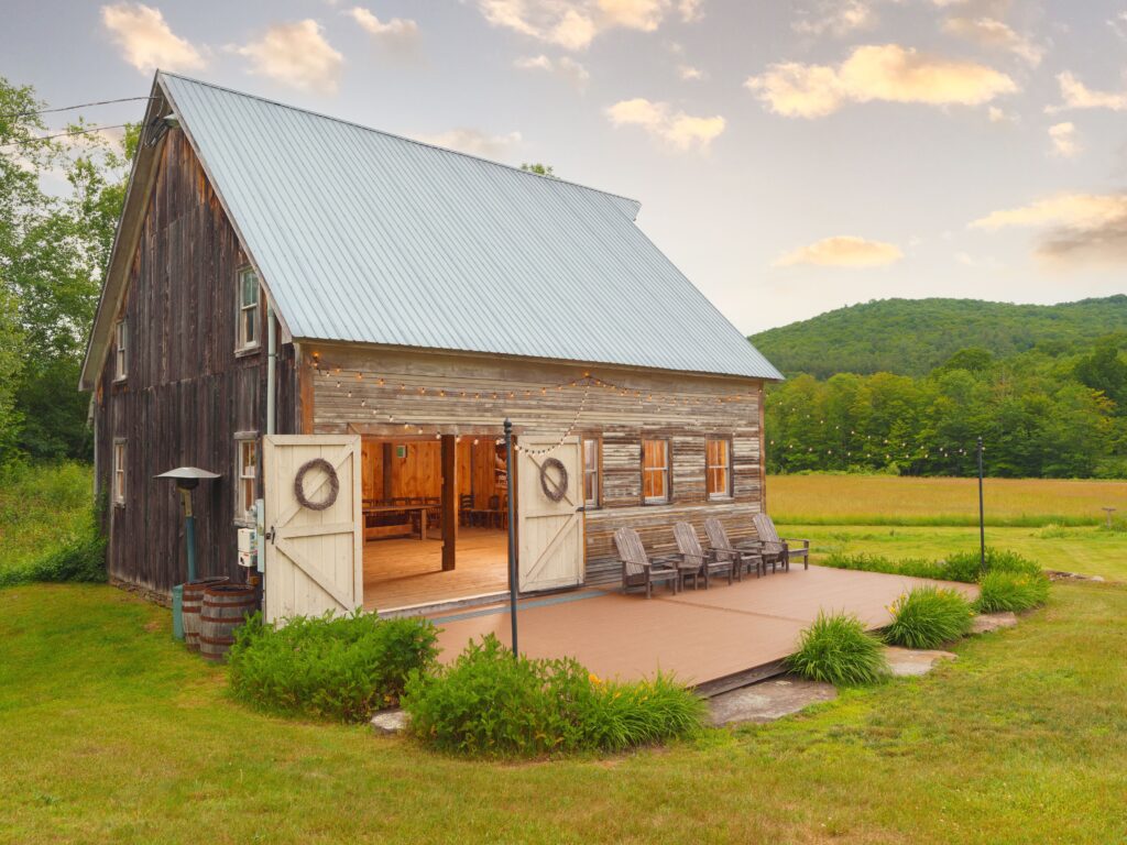 A rustic wood barn with open doors, string lights, and Adirondack chairs on a deck, set against a backdrop of open fields and forested hills at The Farmhouse Inn.